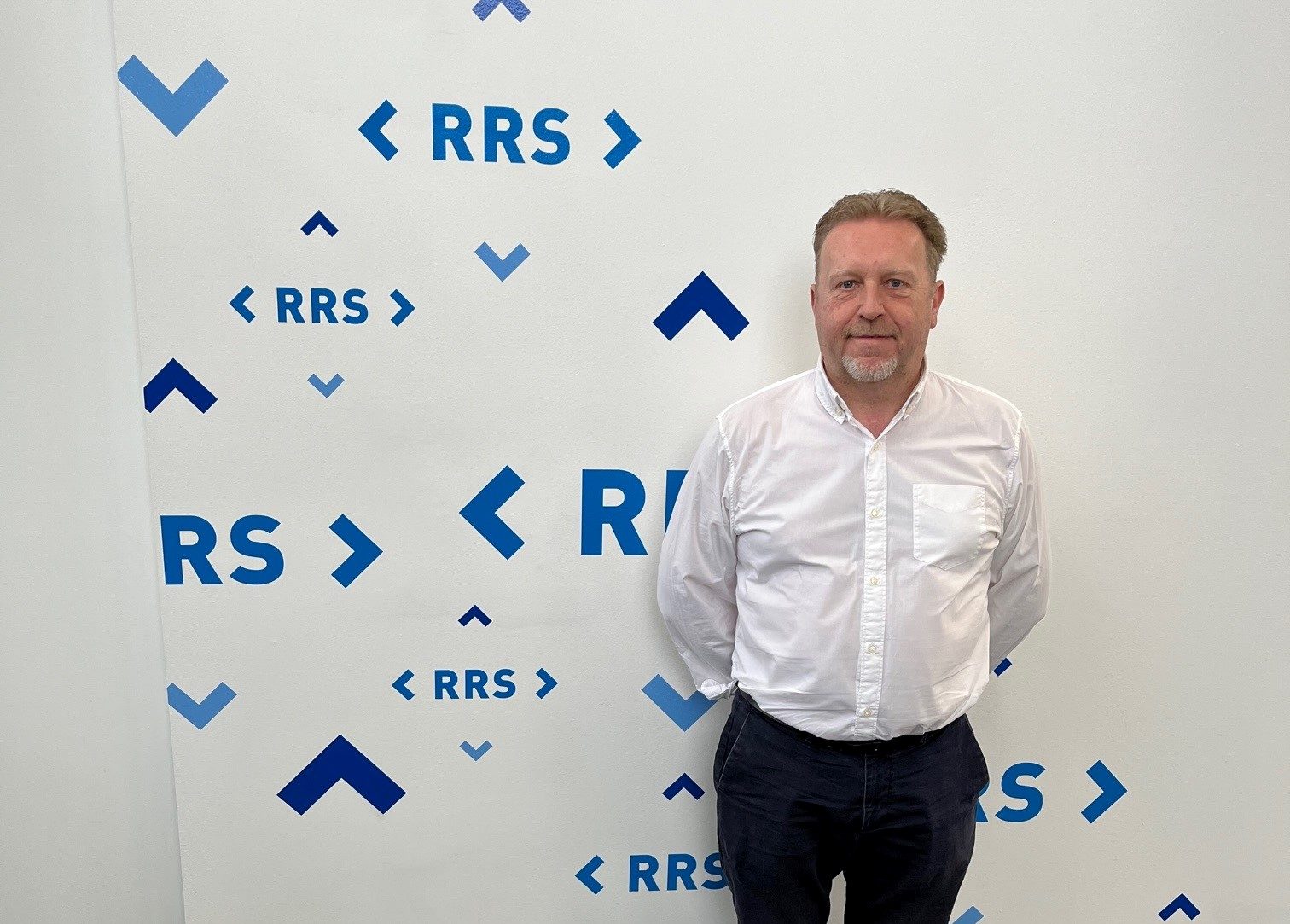 Neil Kennell in white shirt with hands behind back in front of RRS Group branded wall